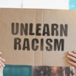 The phrase " Unlearn racism " on a banner in men's hand with blurred background. Stop discrimination. Inequality. Pressure. Tension. Unlawful. Human rights. Illegal. Conflict